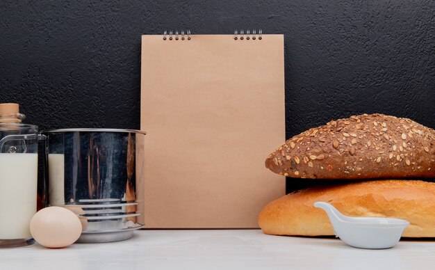 Vue latérale des pains en tant que baguette noire et vietnamienne ensemencée avec tamis à beurre d'oeuf et bloc-notes sur une surface blanche et une surface noire avec espace de copie