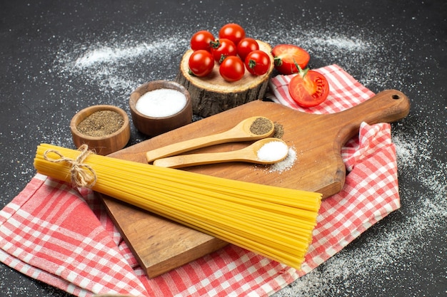 Vue latérale des pâtes crues sel poivre en cuillères sur une planche à découper sur une serviette dépouillée rouge tomates entières coupées sur fond blanc noir