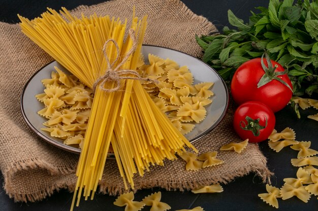 Vue latérale des pâtes crues avec des spaghettis crus sur une assiette avec des tomates et un bouquet de menthe sur une serviette beige