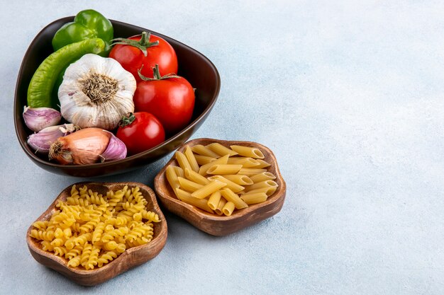 Vue latérale des pâtes non cuites dans des bols avec des tomates ail et poivron dans un bol sur une surface grise