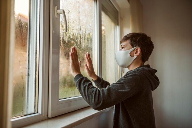 Vue latérale petit garçon avec masque facial regardant à travers la fenêtre