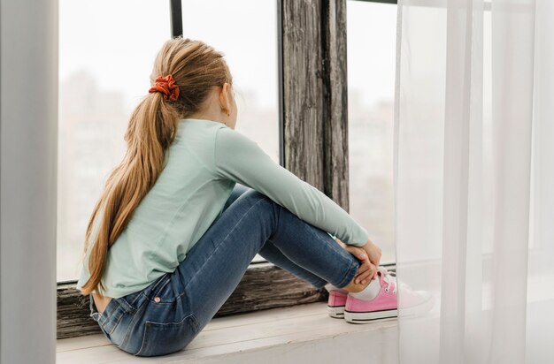 Vue latérale petite fille assise sur un rebord de fenêtre