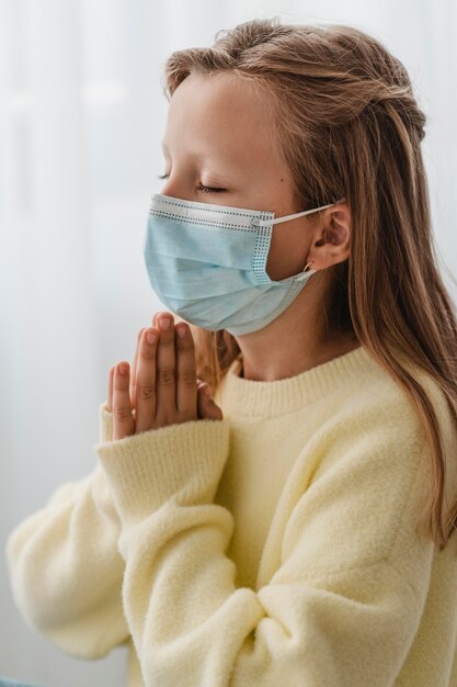 Vue latérale de la petite fille priant avec un masque médical