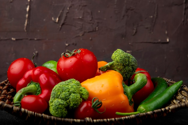 Vue latérale de la plaque de panier pleine de légumes comme le brocoli et les tomates poivrons sur la surface noire et la surface marron
