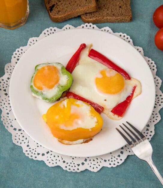 Vue latérale de la plaque de petit déjeuner avec des œufs et des poivrons sur napperon en papier avec une fourchette sur fond bleu