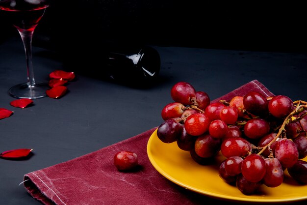 Vue latérale de la plaque avec raisin sur tissu avec verre et bouteille de vin rouge avec des pétales de fleurs sur fond noir