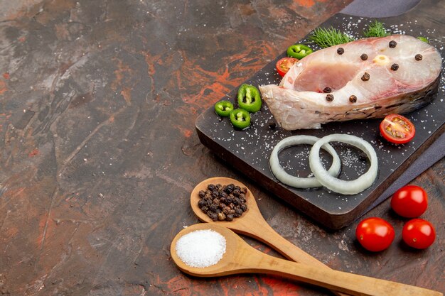 Vue latérale des poissons crus et tomates vertes oignon poivron sur planche à découper noire sur une serviette de couleur foncée sur une surface de couleur mixte