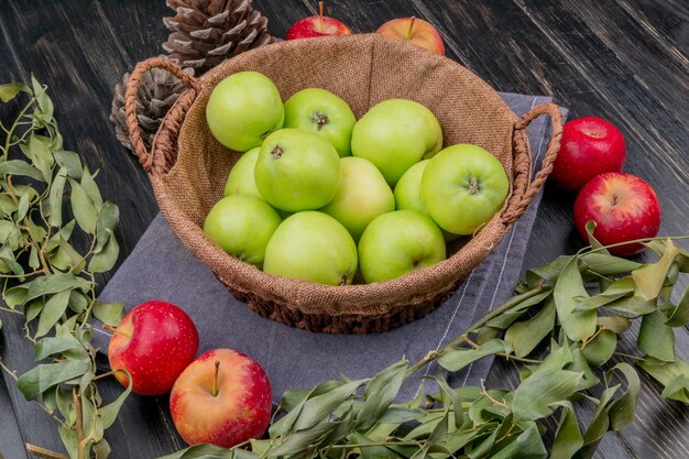 Vue latérale des pommes dans le panier avec des pommes de pin et des feuilles sur tissu et surface en bois