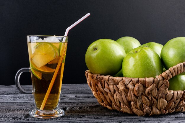 Vue latérale des pommes vertes dans un panier de paille beige et jus sur table en bois gris et noir