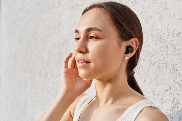 Vue latérale portrait d'une belle femme aux cheveux noirs regardant loin et touchant les airpods, vêtue d'un haut blanc, debout isolée sur fond gris en plein air.