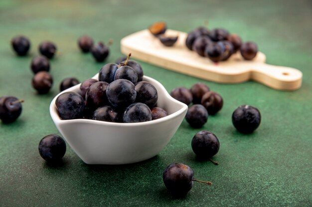 Vue latérale des prunelles violet foncé sur un bol blanc avec prunelles sur planche de cuisine en bois sur fond vert