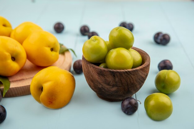 Vue latérale des prunes cerises vertes sur un bol en bois avec des pêches jaunes isolé sur une planche de cuisine en bois sur fond bleu