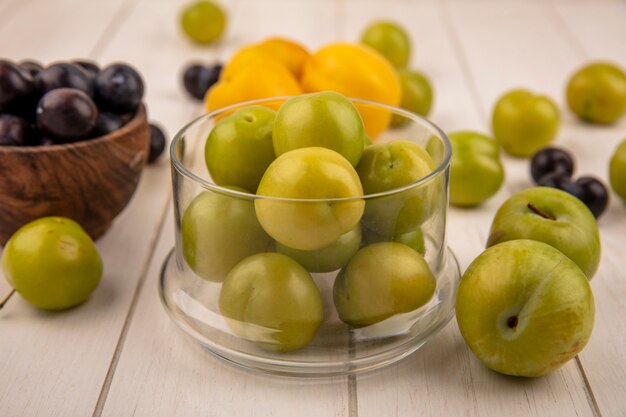 Vue latérale des prunes cerises vertes fraîches sur un bol en verre avec prunelles violet foncé sur un bol en bois avec des pêches isolé sur un fond en bois blanc