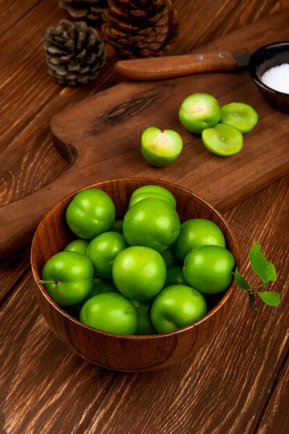 Vue latérale des prunes vertes aigres dans un bol en bois et des prunes en tranches avec un couteau de cuisine et du sel dans une soucoupe sur une table rustique