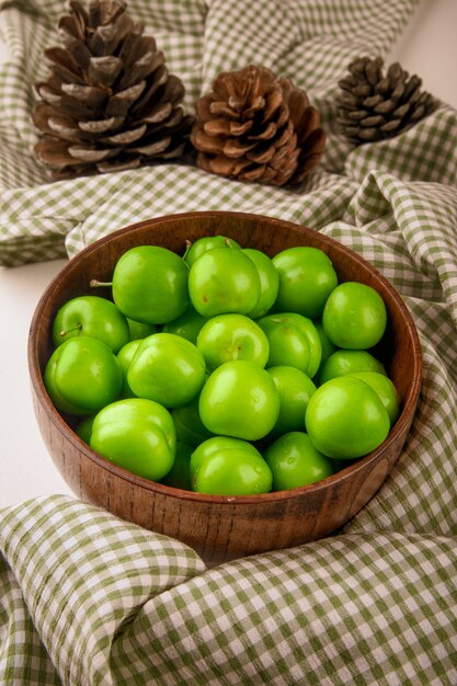 Vue latérale des prunes vertes dans un bol en bois sur une table blanche avec tissu à carreaux
