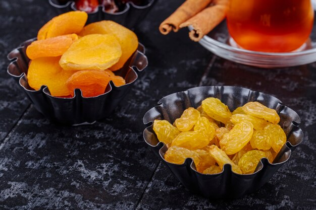 Vue latérale des raisins secs et des abricots secs en mini tartelettes servies avec du thé sur fond de bois noir