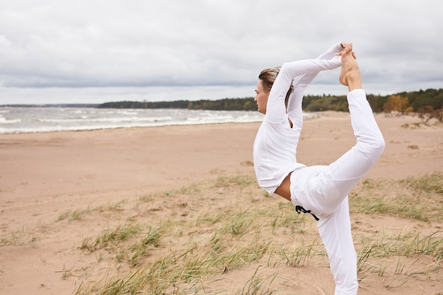 Vue latérale recadrée d'un jeune homme sportif travaillant à l'extérieur, entraînement balanca et flexibilité, faisant du backbend, debout à Natarjasana sur le sable. Yogi masculin maintenant l'équilibre, faisant la pose de danseur King