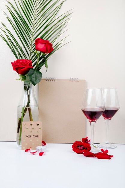 Vue latérale de roses rouges avec feuille de palmier dans une bouteille en verre debout près d'un carnet de croquis et deux verres de vin rouge sur fond blanc