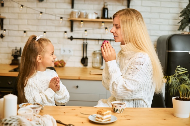 Vue latérale de la séduisante jeune femme de race blanche en pull blanc disant grâce avant le dîner assis à la table de la cuisine avec sa petite fille, en appuyant sur les mains, va manger des gâteaux et boire du thé