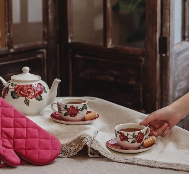 Vue latérale d'un service à thé de motif floral rose avec une théière et une tasse de thé avec des biscuits sur la nappe