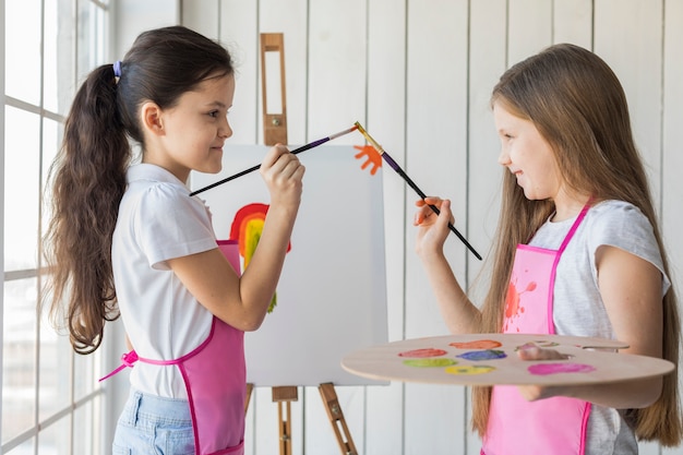 Vue latérale d&#39;un sourire de deux filles touchant leurs pinceaux tout en peignant sur une toile