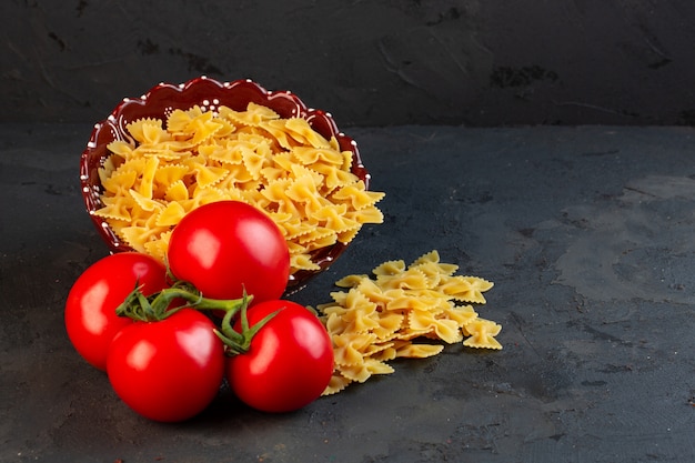 Vue latérale d'un tas de tomates fraîches avec des pâtes crues farfalle éparpillées sur fond noir