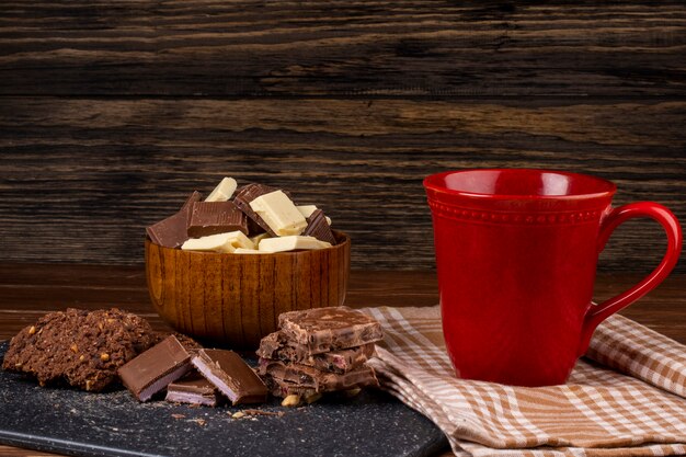 Vue latérale d'une tasse avec des biscuits à l'avoine au thé et des morceaux de chocolat noir et blanc sur fond rustique