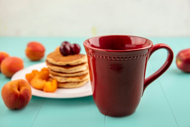 Vue latérale d'une tasse de café avec assiette de crêpes et tranches d'abricot aux cerises sur une surface bleue et fond blanc