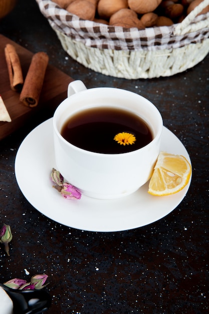 Vue latérale d'une tasse de thé avec une tranche de citron et un bâton de cannelle et un panier avec des noix sur fond noir