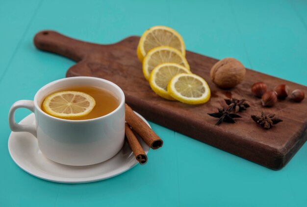 Vue latérale d'une tasse de thé avec tranche de citron et cannelle sur soucoupe avec noix de noix de citron en tranches sur une planche à découper sur fond bleu