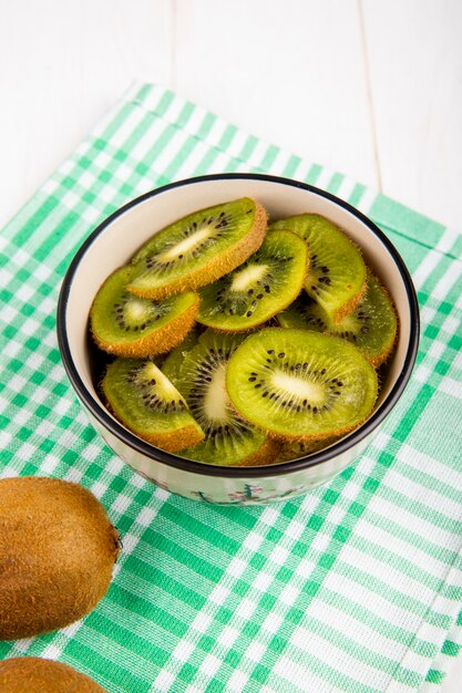 Vue latérale de tranches de kiwi dans un bol sur une serviette de table à carreaux sur blanc