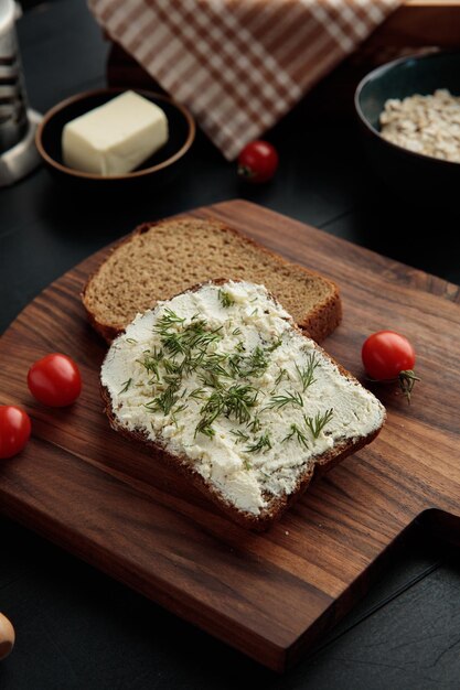 Vue latérale des tranches de pain une enduite de fromage cottage avec de l'aneth dessus et des tomates sur une planche à découper et du beurre avec de l'avoine sur fond noir