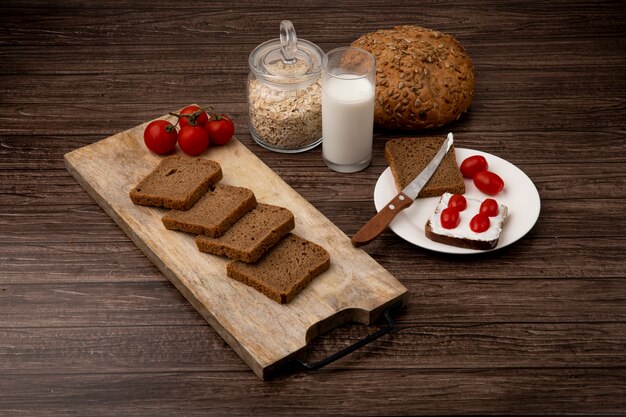 Vue latérale des tranches de pain de seigle et des tomates sur une planche à découper avec des épis de lait et des flocons d'avoine sur fond de bois