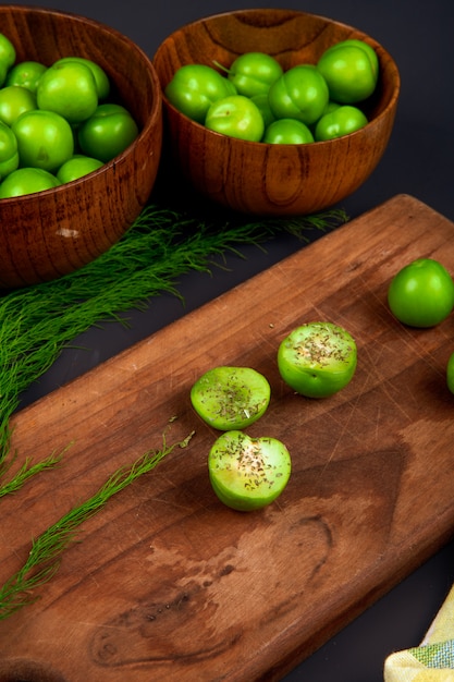 Vue latérale de tranches de prunes vertes saupoudrées de menthe poivrée séchée sur une planche à découper en bois et des bols en bois remplis de prunes vertes sur tableau noir