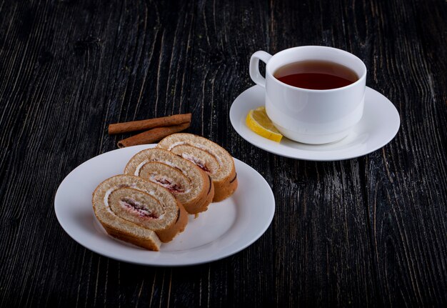 Vue latérale des tranches de rouleau suisse avec de la crème fouettée et de la confiture de framboise sur une assiette servie avec une tasse de thé sur rustique