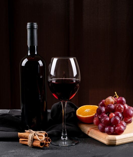 Vue latérale vin rouge avec raisin, orange sur une planche à découper en bois sur une verticale sombre