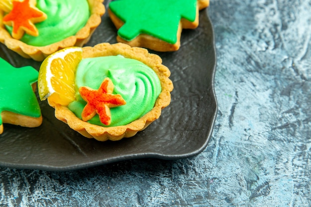 Vue de la moitié inférieure des petites tartes à la crème pâtissière verte biscuits d'arbre de Noël sur une plaque noire sur une surface grise