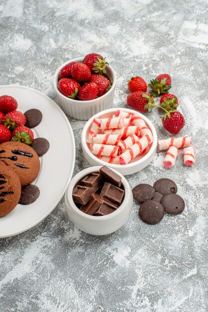 Vue de la moitié supérieure cookies fraises et chocolats ronds sur la plaque ovale blanche et bols de bonbons chocolats fraises sur la table gris-blanc