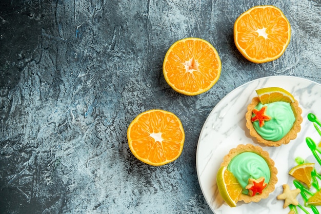 Vue de la moitié supérieure de petites tartelettes à la crème pâtissière verte et tranche de citron sur une plaque ovale oranges coupées sur une surface sombre