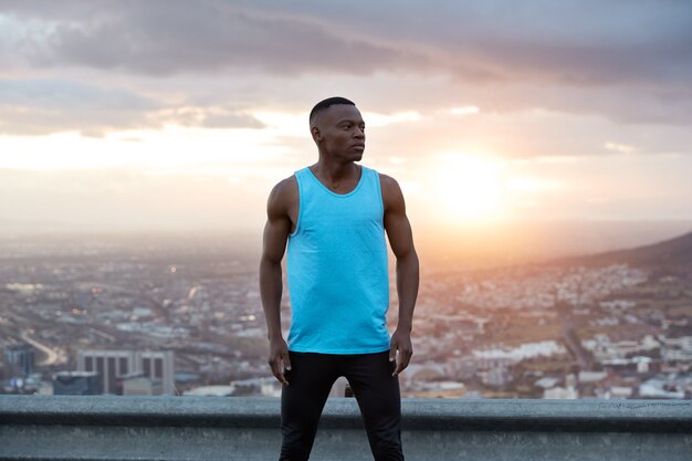 Vue panoramique de l'homme musclé noir gratuit en vêtements de sport, a des exercices en plein air, pose sur un paysage magnifique, lever du soleil à l'aube, bâtiments de la ville, ciel clair, jouit de la liberté, de l'air frais et de la solitude