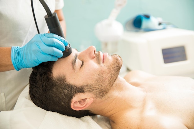 Vue de profil d'un beau jeune homme recevant un traitement facial RF dans un spa de santé