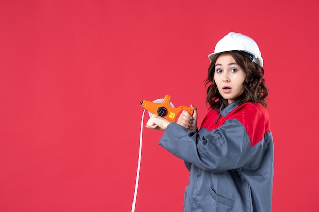 Vue rapprochée de l'architecte femme surprise en uniforme avec un casque à l'aide d'un ruban à mesurer sur mur rouge