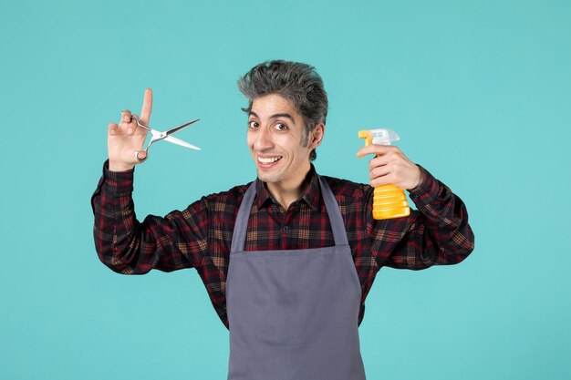 Vue rapprochée avant du barbier masculin heureux et excité portant un tablier gris et tenant un vaporisateur de ciseaux sur un fond de couleur bleu doux