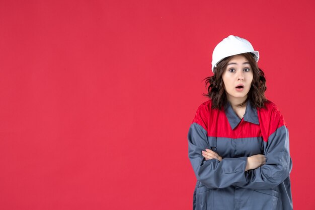 Vue rapprochée avant du constructeur féminin confus en uniforme avec un casque traversant ses bras sur un mur rouge isolé