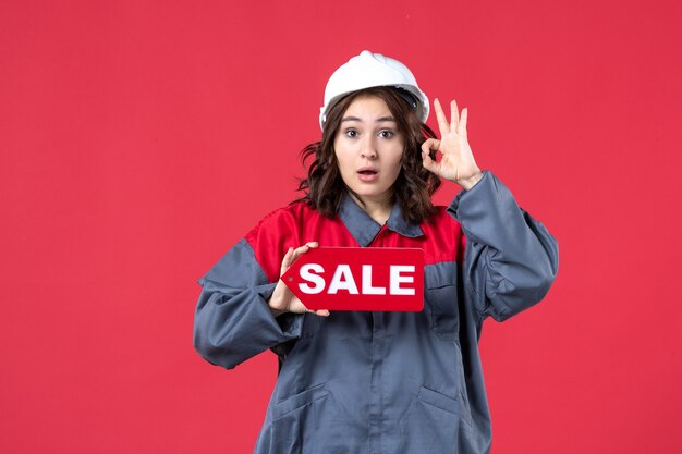 Vue rapprochée avant du travailleur féminin surpris en uniforme portant un casque montrant l'icône de vente et faisant des gestes de lunettes sur un mur rouge isolé