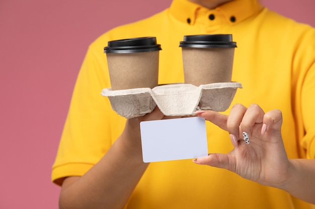 Vue rapprochée avant femme courrier en uniforme jaune cape jaune tenant des tasses à café marron en plastique et une carte sur le bureau rose livraison uniforme couleur féminine