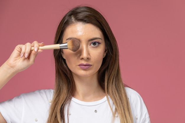 Vue rapprochée avant jeune femme en chemise blanche tenant un pinceau de maquillage sur rose, modèle femme pose fille beauté