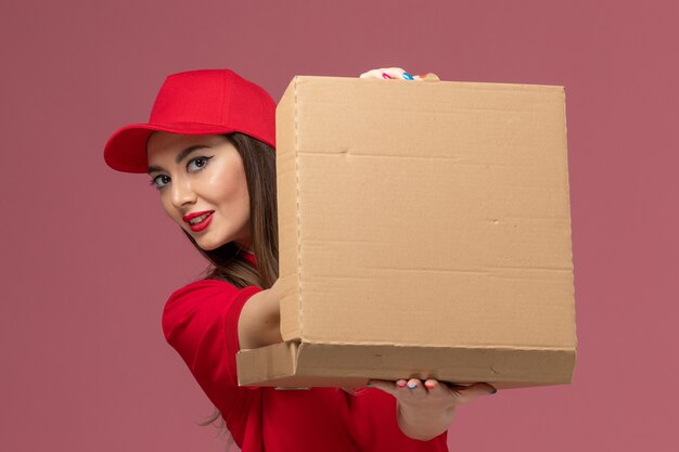 Vue rapprochée avant jeune femme courrier en uniforme rouge tenant la boîte de nourriture de livraison sur fond rose clair service de livraison de travail entreprise uniforme