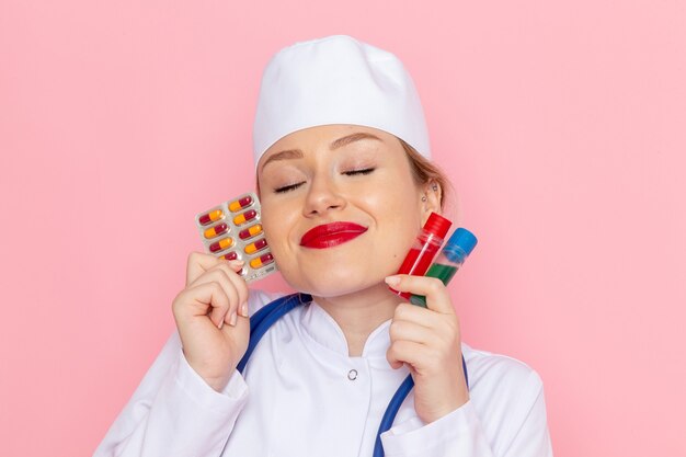 Vue rapprochée avant jeune femme médecin en costume médical blanc avec stéthoscope bleu tenant des pilules et des flacons sur rose