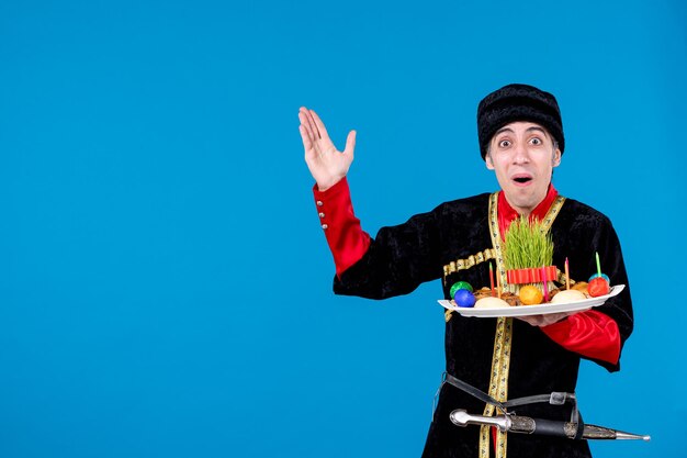 Vue rapprochée avant d'un jeune homme choqué en costume traditionnel tenant un plateau rempli de confiseries nationales et pointant vers le haut sur le côté droit sur fond bleu